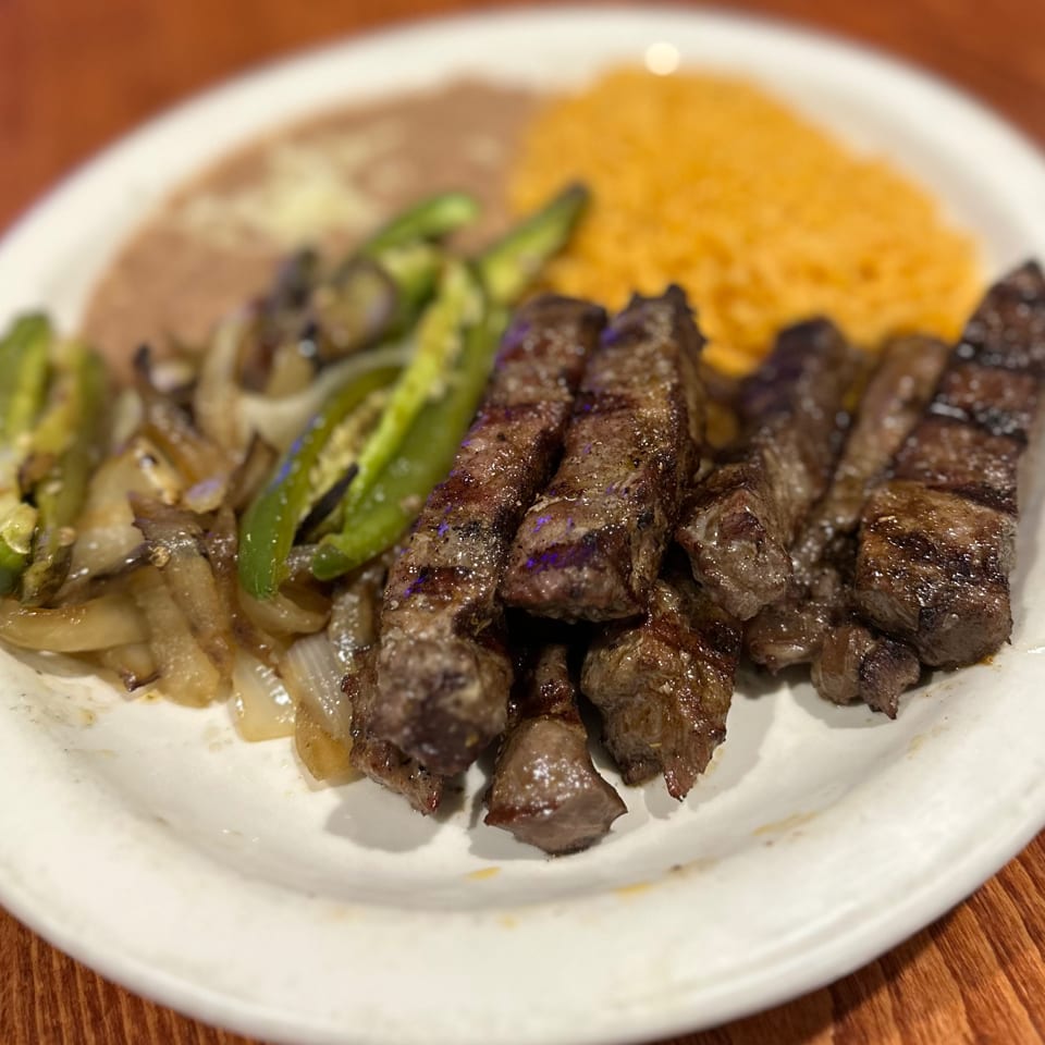 Grilled steak with sautéed onions, green peppers, refried beans, and yellow rice on a white plate.
