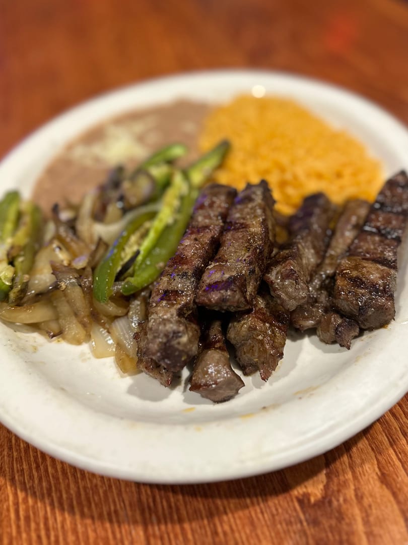 Grilled steak with sautéed onions, green peppers, refried beans, and yellow rice on a white plate.