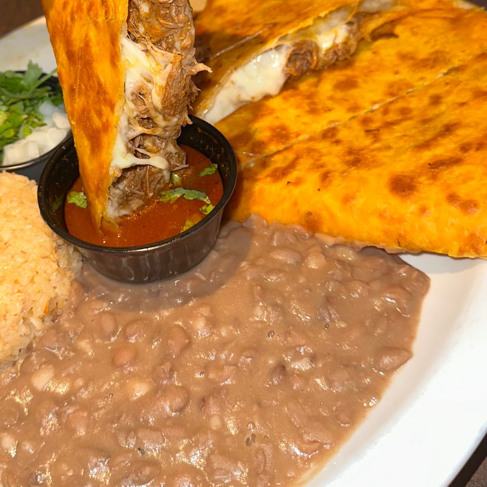 Hand dipping a cheesy birria taco into a small bowl of consommé, with rice and refried beans.