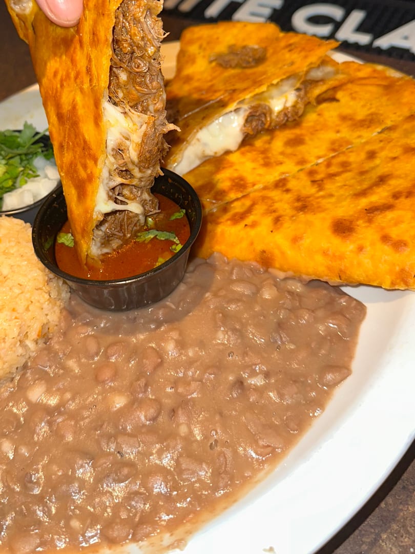 Hand dipping a cheesy birria taco into a small bowl of consommé, with rice and refried beans.