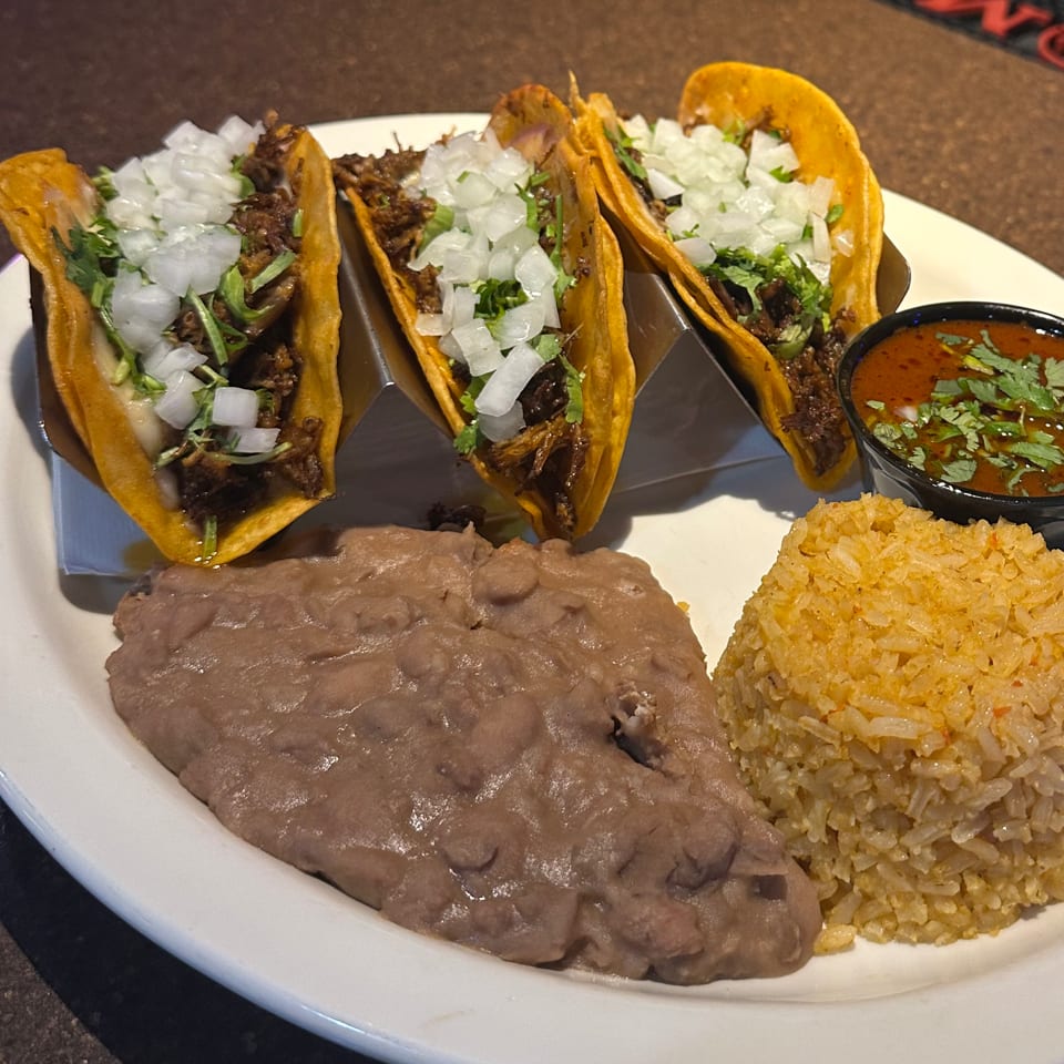 Three Johnny Gringo's Mexican street tacos with rice, refried beans, and a side of red sauce.