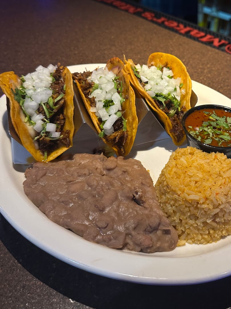 Three Johnny Gringo's Mexican street tacos with rice, refried beans, and a side of red sauce.