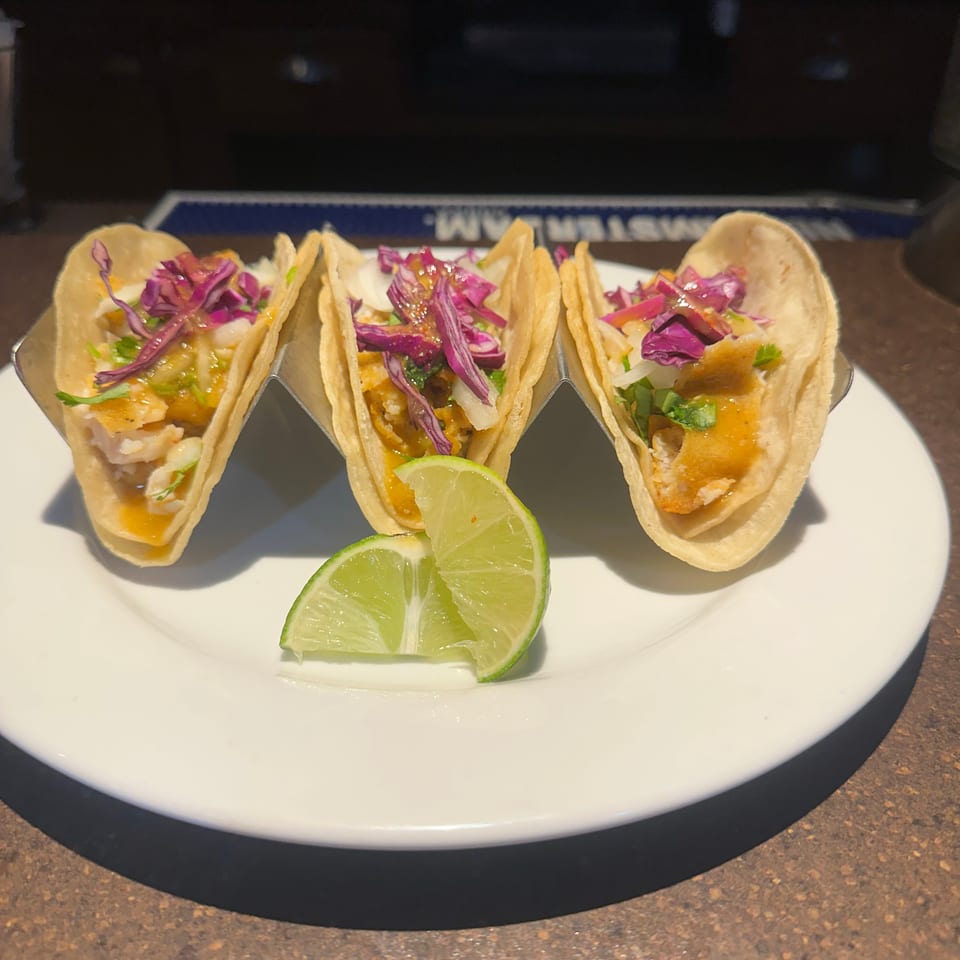 Three fish tacos with red cabbage, sauce, and cilantro, served with lime wedges on a white plate.